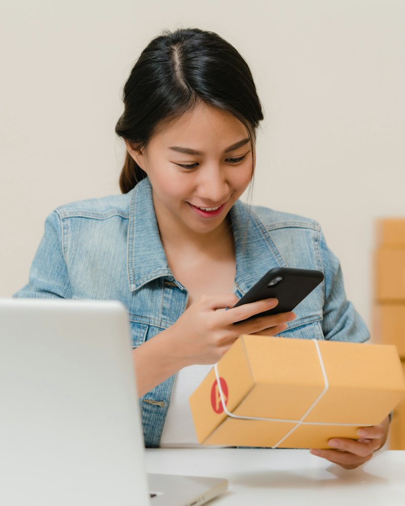Eine Person am Schreibtisch hält ein Paket und ein Telefon, daneben ein Laptop